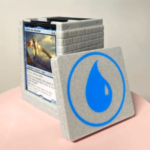 May include: A gray, brick-textured card storage box with a lid featuring a blue water drop symbol. The box holds several trading cards, one of which is visible, displaying the text "Kefnet the Mindful". The box is on a pink surface.