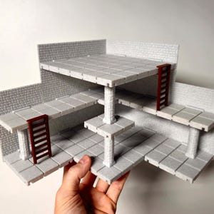 May include: A multi-level, gray miniature dungeon terrain set with brick walls, tiled floors, and brown ladders. The set is held by a hand and features multiple platforms supported by pillars, creating a three-dimensional structure for tabletop gaming.