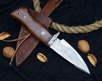 May include: A brown wooden handled knife with a Damascus steel blade. The knife is in a brown leather sheath with a decorative stitching pattern. The knife is on a black fabric background with a rope and nuts.