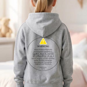 Może przedstawiać: Szara bluza z kapturem z okrągłą grafiką na plecach. Grafika zawiera żółty znak ostrzegawczy i tekst w języku niemieckim. Tekst ostrzega przed bardzo zmotywowaną zawodniczką piłki ręcznej. Bluza jest noszona przez osobę.