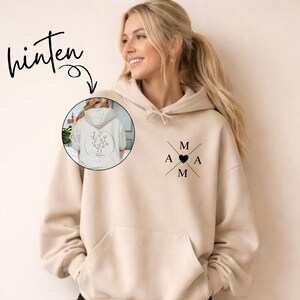 May include: A beige hoodie with a front pocket and a hood. The front features a black heart and the letters "M A M" in a cross design. The back of the hoodie, shown in a circular inset, displays a floral design within a dotted circle.
