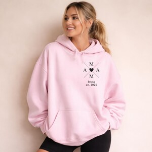 May include: A light pink hoodie with a front pocket and a hood. The hoodie features a black graphic design on the chest with the letters "M A A M" arranged in a cross shape, a heart, and the text "Emma est. 2025".
