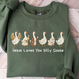Puede incluir: Sudadera verde militar con el texto "Jesus Loves You Silly Goose". La sudadera presenta un gráfico de gansos vestidos con atuendo religioso. Los gansos son blancos con picos naranjas y visten túnicas y sostienen bastones.