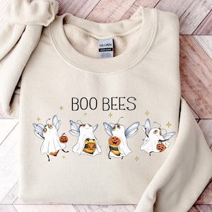 Può includere: Felpa color sabbia con la scritta "BOO BEES" e una grafica di api vestite da fantasmi, che tengono zucche e secchielli di caramelle. Il design include piccole stelle. Un top a maniche lunghe per Halloween.