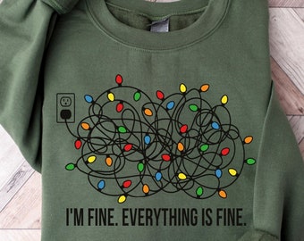 Puede incluir: Una sudadera de cuello redondo color verde militar con un gráfico de un cable eléctrico negro enredado con bombillas navideñas de colores conectado a un enchufe. El texto debajo del gráfico dice, I'M FINE. EVERYTHING IS FINE.
