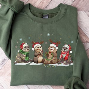 Puede incluir: Sudadera verde militar con un diseño festivo de cuatro nutrias con gorros de Papá Noel, bufandas y cuernos de reno. Las nutrias están rodeadas de luces navideñas y copos de nieve, creando un tema navideño.