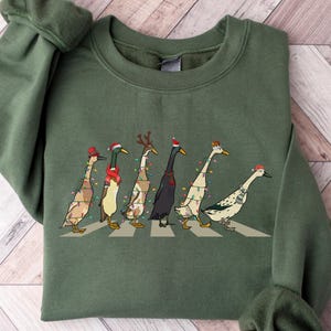 Peut inclure: Un sweat-shirt vert avec un motif de No&euml;l repr&eacute;sentant une ligne de canards portant des chapeaux de P&egrave;re No&euml;l et des lumi&egrave;res de No&euml;l. Le texte "Military Green" est affich&eacute; en haut de l'image.