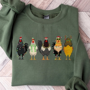 May include: A military green crewneck sweatshirt featuring a graphic of five chickens decorated for the holidays. The chickens are wearing festive accessories including Santa hats, a green and white scarf, reindeer antlers, and colorful string lights.