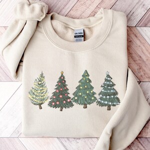 Könnte beinhalten: Ein sandfarbenes Sweatshirt mit Rundhalsausschnitt und einem Grafikdruck von vier stilisierten Weihnachtsbäumen. Jeder Baum ist mit verschiedenfarbigen Lichtern in Gelb, Rot und Weiß geschmückt, und ein Baum ist mit einem kleinen gelben Stern versehen.