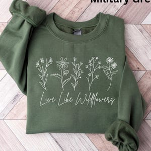 Puede incluir: Sudadera verde militar con diseño floral blanco y el texto "Live Like Wildflowers". La sudadera de cuello redondo tiene mangas con puños. La prenda se exhibe sobre una superficie de madera. La sudadera está hecha de un material suave.