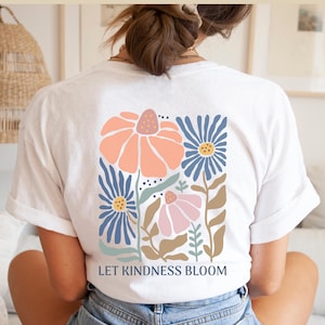 May include: White t-shirt featuring a floral design in peach, blue, and pink. The graphic shows stylized flowers and the text "LET KINDNESS BLOOM". The shirt is made of a soft material. The design is on the back of the shirt.