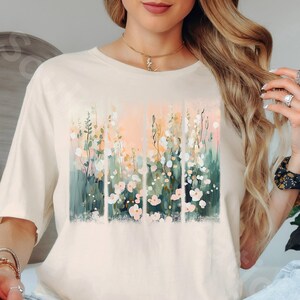 May include: Cream-colored t-shirt featuring a floral design. The artwork showcases a vertical arrangement of flowers in shades of pink, white, green, and peach. The shirt has a classic crew neck and short sleeves, suitable for casual wear.