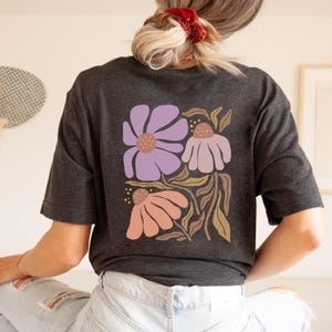 May include: A dark charcoal gray short-sleeve t-shirt featuring a large graphic on the back. The design displays three stylized flowers in shades of lavender, light pink, and peach, accompanied by tan-colored stems and leaves on a dark background.