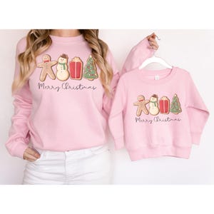 May include: Two pink sweatshirts with a gingerbread man, snowman, gift, and Christmas tree design. The text "Merry Christmas" is below the graphic. One sweatshirt is adult-sized, and the other is child-sized.