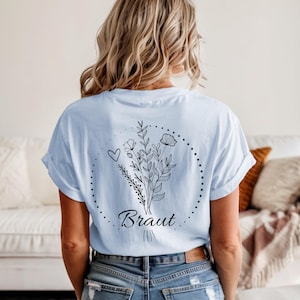 Puede incluir: Camiseta azul claro con un dise&ntilde;o floral y la palabra "Braut" en cursiva. El dise&ntilde;o incluye un coraz&oacute;n, flores y hojas dentro de un c&iacute;rculo punteado. La camiseta es de estilo casual.