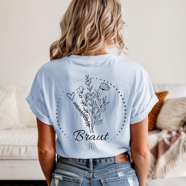 Puede incluir: Camiseta azul claro con un dise&ntilde;o floral y la palabra "Braut" en cursiva. El dise&ntilde;o incluye un coraz&oacute;n, flores y hojas dentro de un c&iacute;rculo punteado. La camiseta es de estilo casual.