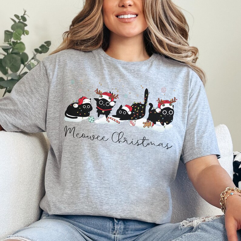 May include: A light gray t-shirt featuring a festive design of five black cats wearing Santa hats and reindeer antlers. The cats are decorated with Christmas lights and candy canes, with the words "Meowee Christmas" in script.