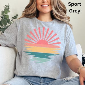 May include: Sport Grey t-shirt featuring a colorful sunset design. The graphic includes a pink sun with orange, yellow, and teal brushstrokes. The text "Sport Grey" is printed in black on the upper right side of the shirt.