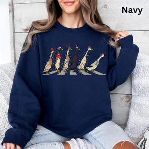 Peut inclure: Un sweat-shirt bleu marine avec un motif de canards portant des lumi&egrave;res de No&euml;l et des chapeaux marchant sur un passage pour pi&eacute;tons. Les canards portent des chapeaux de P&egrave;re No&euml;l, des bois de renne et une vari&eacute;t&eacute; d'autres accessoires festifs.