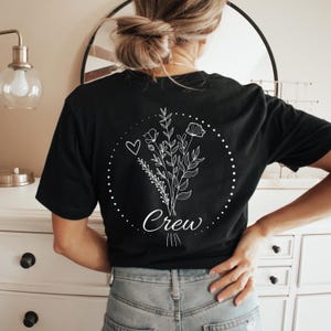 May include: Black t-shirt with a white floral design and the word "Crew" in script. The design includes a heart and a dotted circle around the flowers. The shirt is made of a soft material.