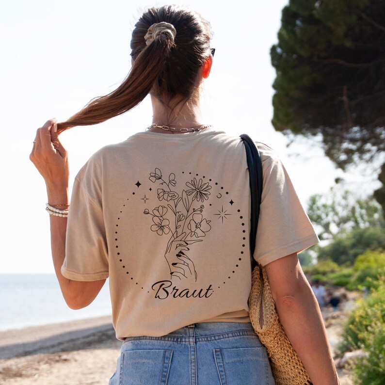 Pu&ograve; includere: T-shirt beige con un design floreale nero e la parola "Braut" in corsivo. Il design presenta una mano che tiene un bouquet di fiori all'interno di un cerchio tratteggiato, con stelle e altre forme. La maglietta &egrave; abbinata a jeans blu.