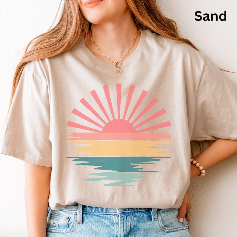 May include: A sand-colored t-shirt featuring a colorful sunset design. The graphic includes a pink sun with radiating lines, a yellow and pink horizon, and a teal water reflection. The word "Sand" is printed in black.