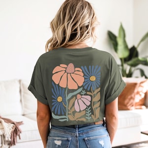 May include: Olive green t-shirt featuring a floral design on the back. The design includes large orange, blue, and pink flowers with green and brown leaves. The shirt has rolled-up sleeves and is paired with blue jeans.