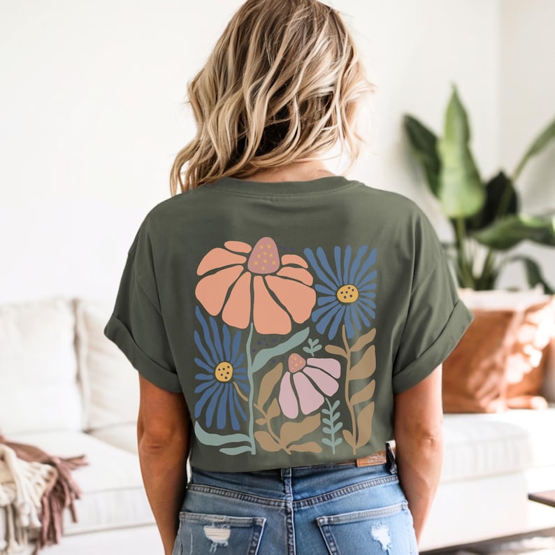 May include: Olive green t-shirt featuring a floral design on the back. The design includes large orange, blue, and pink flowers with green and brown leaves. The shirt has rolled-up sleeves and is paired with blue jeans.
