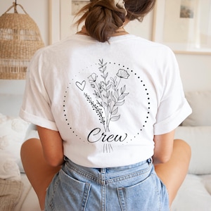 May include: White t-shirt with a black floral design and the word "Crew" in script. The design includes a heart and a dotted circle. The shirt is made of a soft material and is suitable for casual wear.
