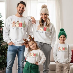 May include: White sweatshirts with a gingerbread man, snowman, gift, and Christmas tree design. The text "Merry Christmas" is below the graphic. The family is wearing matching sweatshirts. The little girl is wearing a green skirt and reindeer antlers.