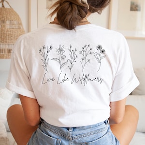 Könnte beinhalten: Weißes T-Shirt mit schwarzer Strichzeichnung von Wildblumen und dem Schriftzug "Live Like Wildflowers". Das florale Design befindet sich auf der Rückseite des Shirts. Das Shirt ist aus weichem, hellem Stoff.
