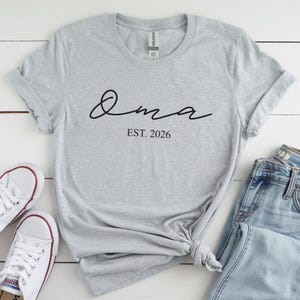 May include: A light gray t-shirt with the word "Oma" in black script and the text "EST. 2026" below it. The shirt is styled with rolled-up sleeves and a knot at the bottom. White sneakers and blue jeans are visible.