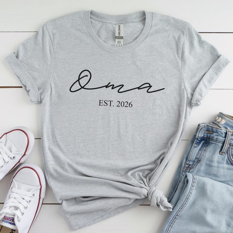 May include: A light gray t-shirt with the word "Oma" in black script and the text "EST. 2026" below it. The shirt is styled with rolled-up sleeves and a knot at the bottom. White sneakers and blue jeans are visible.