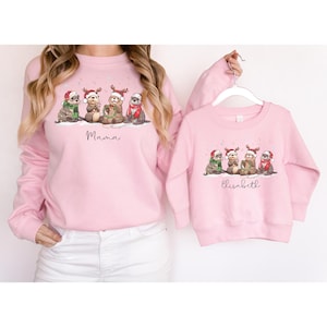May include: Pink sweatshirts with a Christmas otter design. The adult sweatshirt says "Mama" and the child's sweatshirt says "Elisabeth". The otters are wearing Santa hats and scarves. A matching set for mother and child.