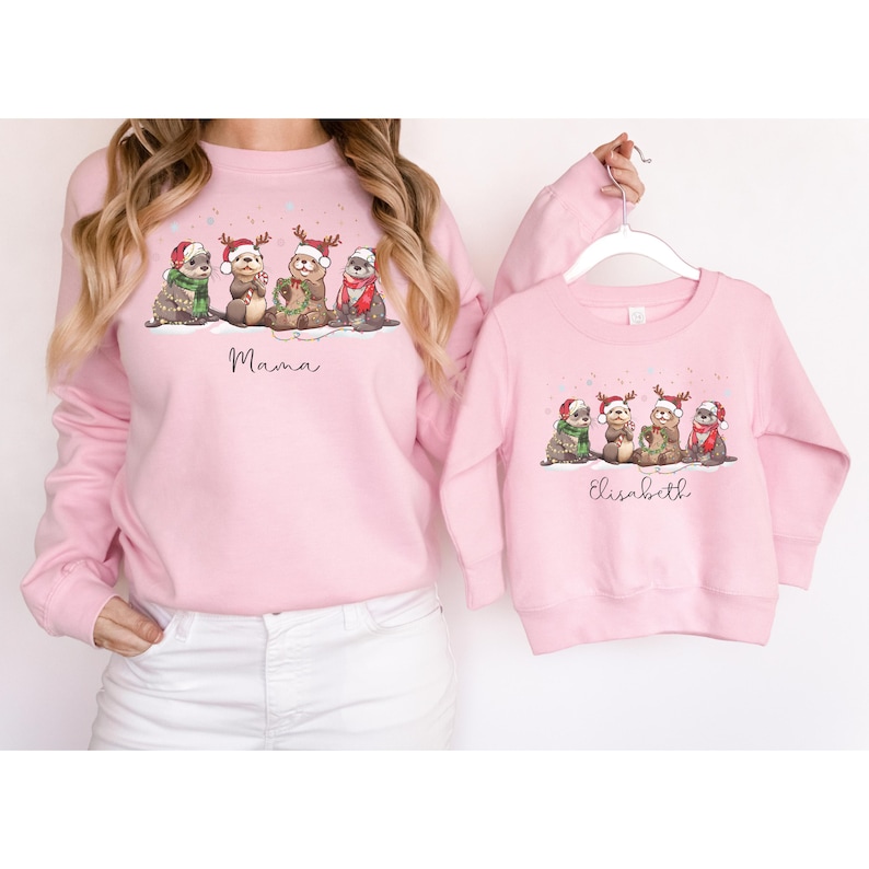 May include: Pink sweatshirts with a Christmas otter design. The adult sweatshirt says "Mama" and the child's sweatshirt says "Elisabeth". The otters are wearing Santa hats and scarves. A matching set for mother and child.