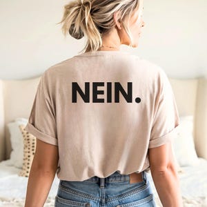 May include: A beige t-shirt with the word "NEIN." printed in bold black letters. The shirt has rolled-up sleeves and is worn with blue jeans. The person is facing away from the camera.