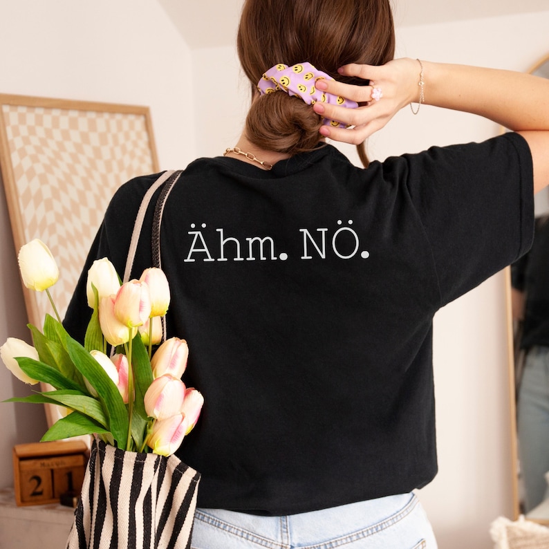 以下が含まれることがあります： 白文字で「&Auml;hm. N&Ouml;.」と書かれた黒いTシャツ。黄色いスマイリーフェイスのシュシュを着用。白黒ストライプのトートバッグには、チューリップのブーケが入っています。
