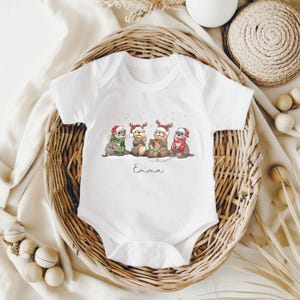 May include: White baby onesie featuring a whimsical illustration of four otters wearing Santa hats and scarves, with the name "Emma" printed below the illustration. The otters are depicted in a winter scene.