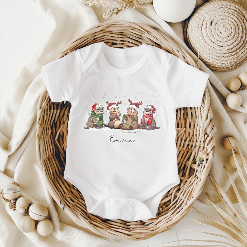 May include: White baby onesie featuring a whimsical illustration of four otters wearing Santa hats and scarves, with the name "Emma" printed below the illustration. The otters are depicted in a winter scene.