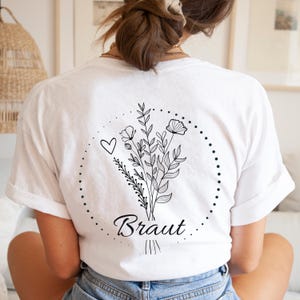 Puede incluir: Camiseta blanca con un dise&ntilde;o de ramo floral negro, un coraz&oacute;n y la palabra "Braut" en cursiva. El ramo est&aacute; rodeado por una l&iacute;nea punteada. La camiseta tiene mangas cortas.