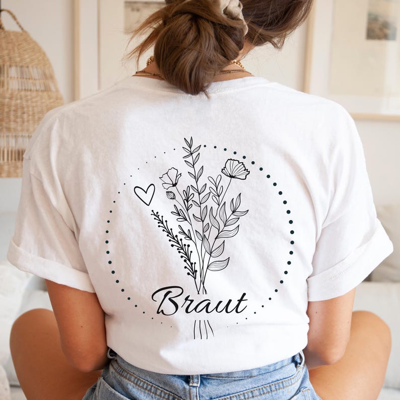 Puede incluir: Camiseta blanca con un dise&ntilde;o de ramo floral negro, un coraz&oacute;n y la palabra "Braut" en cursiva. El ramo est&aacute; rodeado por una l&iacute;nea punteada. La camiseta tiene mangas cortas.