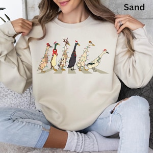 Peut inclure: Un sweat-shirt beige avec un motif de canards portant des chapeaux de No&euml;l et des lumi&egrave;res, traversant un passage pour pi&eacute;tons. Le texte "Sand" est dans le coin sup&eacute;rieur droit de l'image.