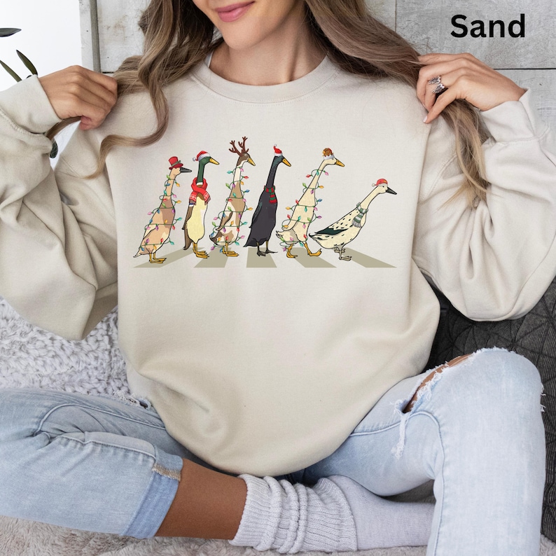 Peut inclure: Un sweat-shirt beige avec un motif de canards portant des chapeaux de No&euml;l et des lumi&egrave;res, traversant un passage pour pi&eacute;tons. Le texte "Sand" est dans le coin sup&eacute;rieur droit de l'image.