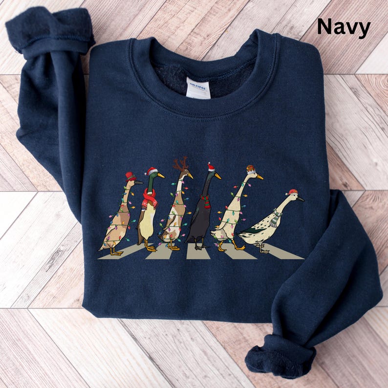 Peut inclure: Sweat-shirt bleu marine avec un motif de sept canards portant des lumi&egrave;res de No&euml;l et traversant un passage pour pi&eacute;tons. Les canards portent des bonnets de P&egrave;re No&euml;l et l'un d'eux a un bandeau de renne.