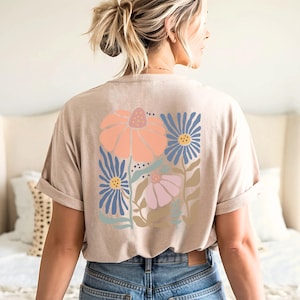 May include: Beige t-shirt with a floral design on the back. The design features a large peach-colored flower, blue flowers, and a pink flower with green leaves. The shirt has rolled-up sleeves and is worn with blue jeans.