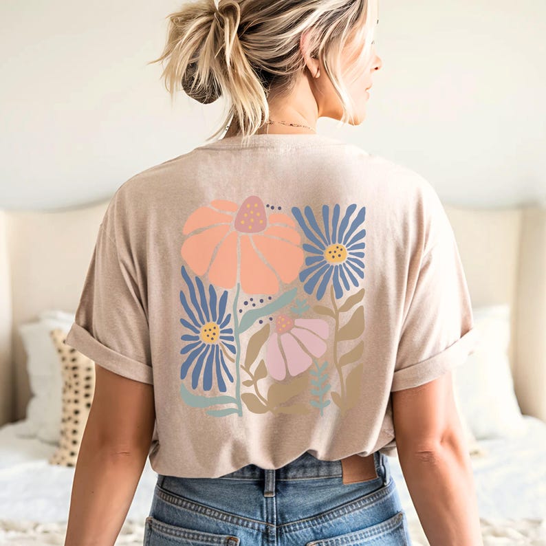 May include: Beige t-shirt with a floral design on the back. The design features a large peach-colored flower, blue flowers, and a pink flower with green leaves. The shirt has rolled-up sleeves and is worn with blue jeans.