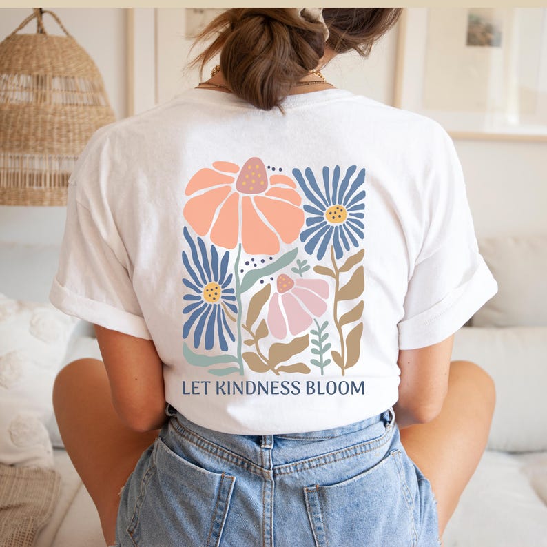May include: White t-shirt featuring a floral design in peach, blue, and pink. The graphic shows stylized flowers and the text "LET KINDNESS BLOOM". The shirt is made of a soft material. The design is on the back of the shirt.
