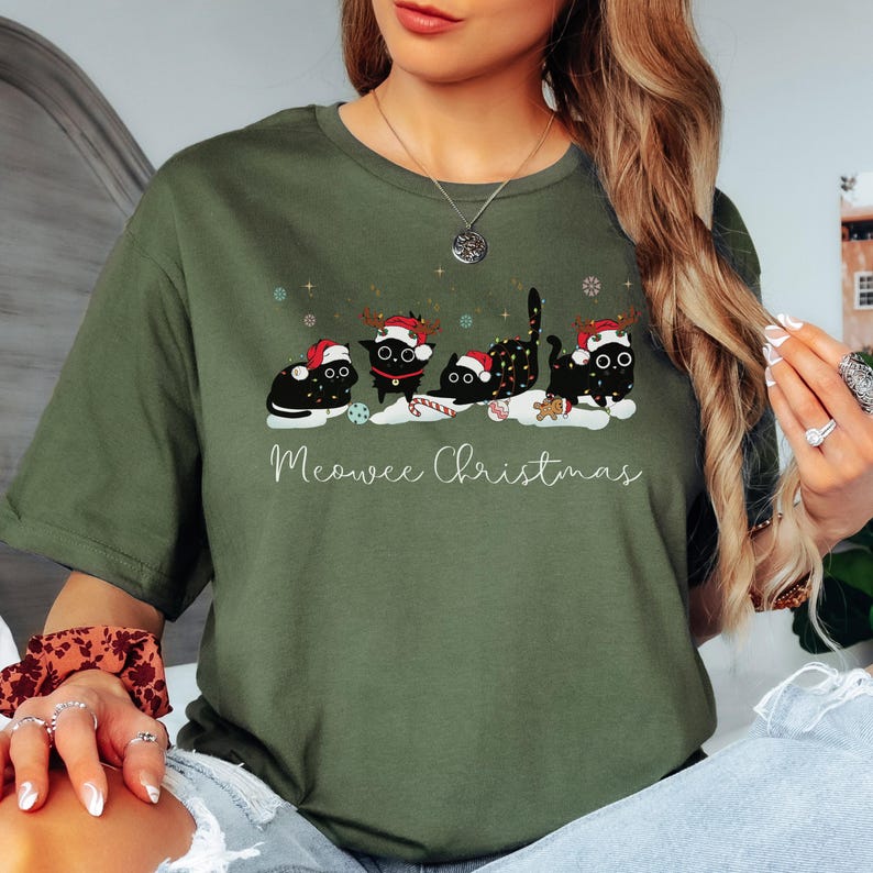 May include: An olive green t-shirt featuring a festive design with four black cats wearing Santa hats and reindeer antlers. The cats are arranged in a row with snowflakes and the text "Meowee Christmas".