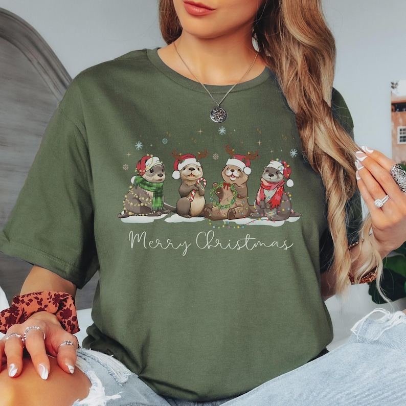May include: An olive green crew neck t-shirt featuring a graphic of four cartoon otters wearing Santa hats and winter scarves. The otters are decorated with string lights and hold holiday items. The text Merry Christmas is printed below in white.