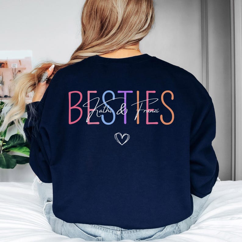 May include: A navy blue sweatshirt featuring the word "BESTIES" in a rainbow of colours. Below, in a script font, are the names "Kathie & Franzi", with a small heart symbol beneath the names.